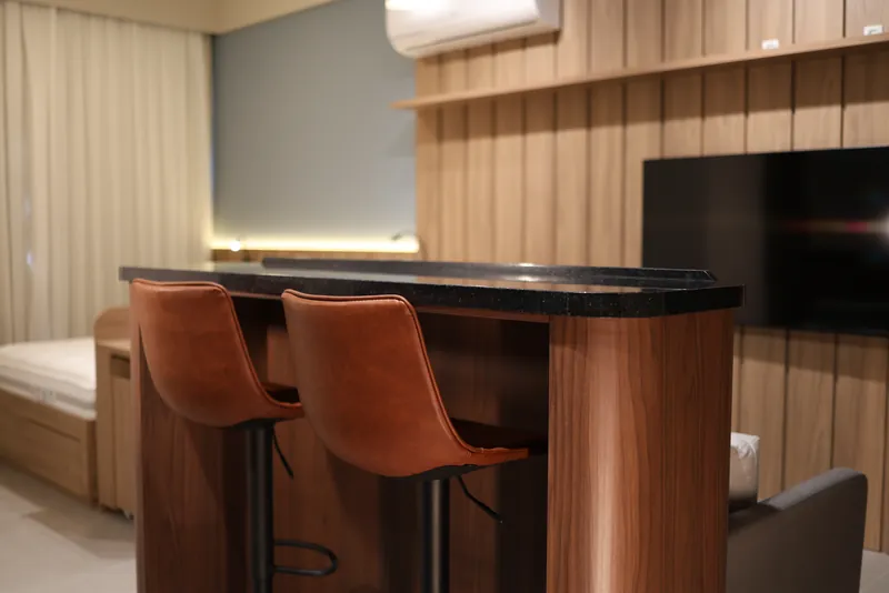 Bancada alta de madeira com duas banquetas de couro caramelo, diante do painel ripado.