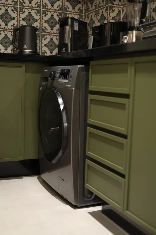 Máquina de lavar embutida em armário verde, com bancada e azulejo estampado acima.